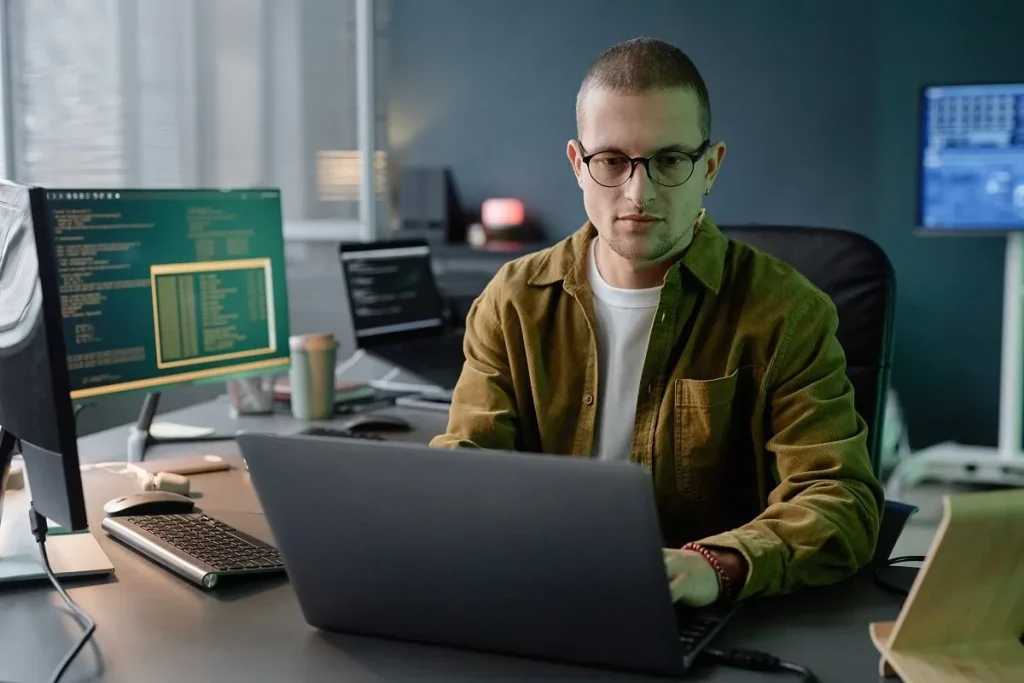 A computer systems analyst working on a laptop in an office with multiple monitors